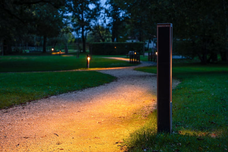 NOWATT solar bollard softly illuminating a gravel pathway at dusk, enhancing visibility and ambiance in a landscaped park setting with off-grid architectural lighting.