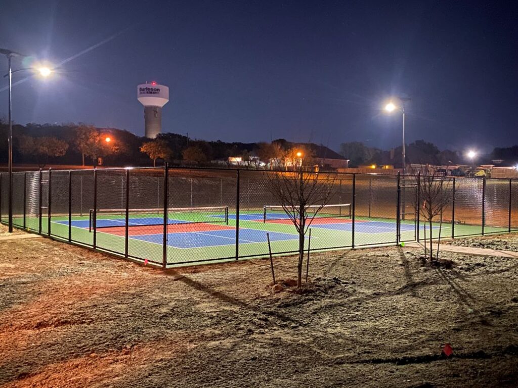 Well-lit outdoor pickleball courts at night illuminated by off-grid solar lighting, with a water tower and neighborhood in the background.