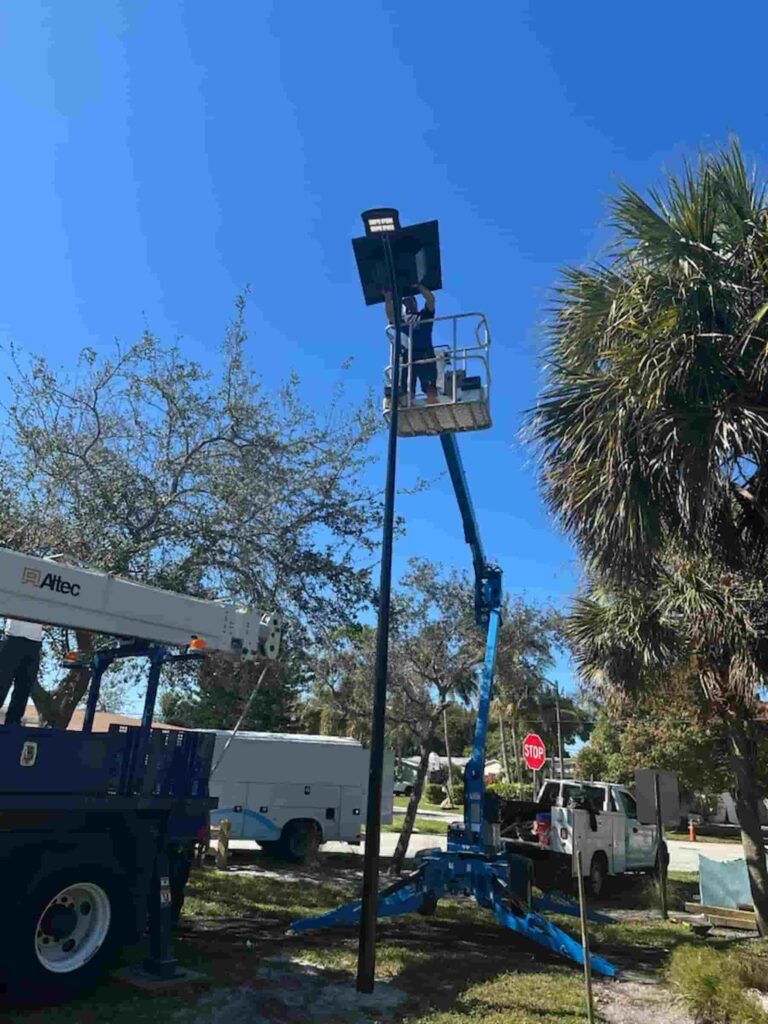 Technician installing a solar-powered streetlight on a flood-prone county road using a lift truck, surrounded by palm trees and maintenance vehicles under a clear blue sky.