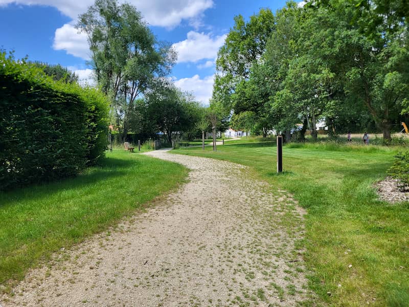 NOWATT solar bollard installed along a sunlit park path, providing clean, off-grid lighting in a green public space with trees and open sky.