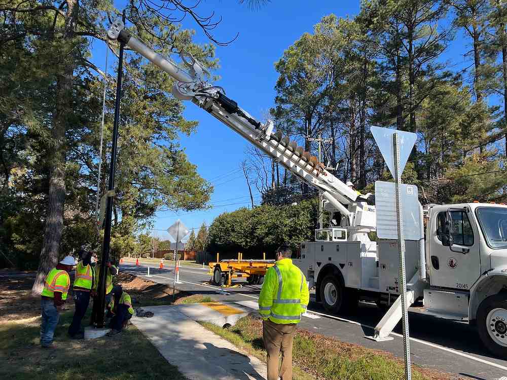 Utility crew installing a light pole with a digger truck along a roadside, representing roadway infrastructure deployment.