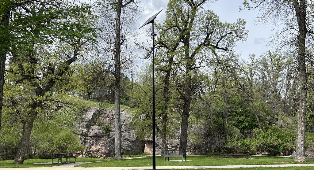 Fonroche solar streetlight installed along a paved path in a wooded public park with rock outcroppings and trees.