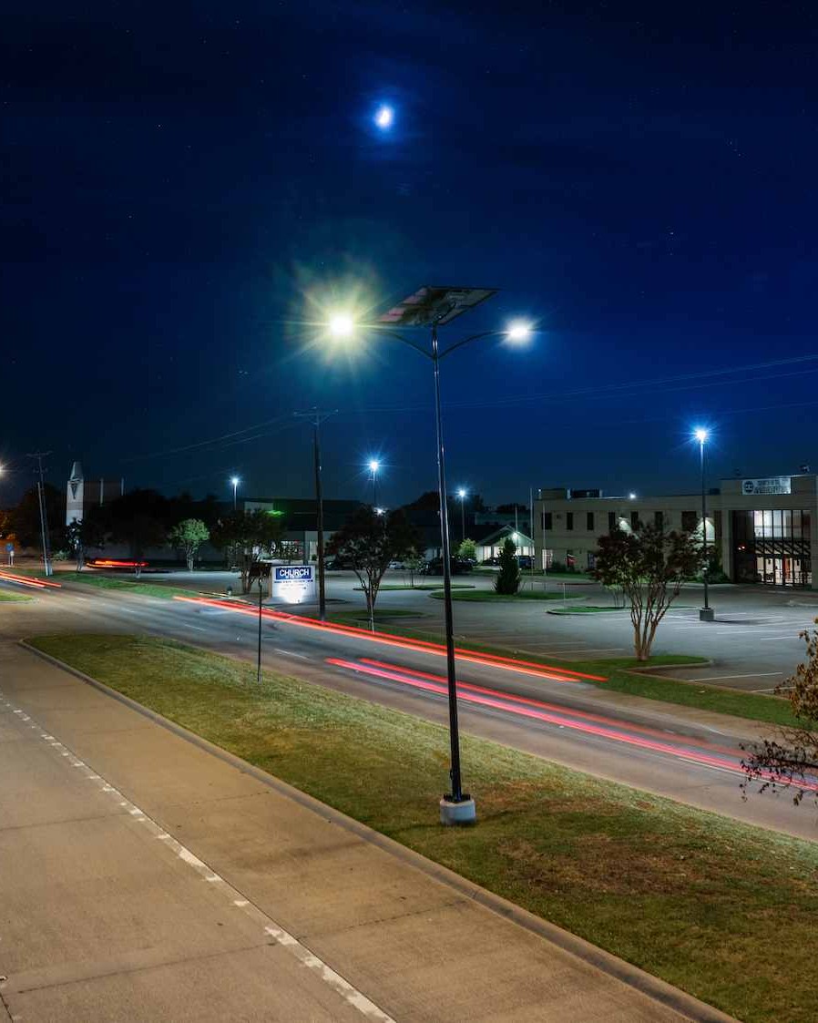 Night view of a solar-powered LED streetlight illuminating Route 66 in Rowlett, Texas, with light trails from passing vehicles and nearby buildings in the background.