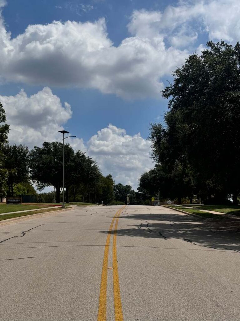 Daytime view of a quiet suburban road in Alabama with solar-powered streetlights and tree-lined sidewalks under a partly cloudy sky.