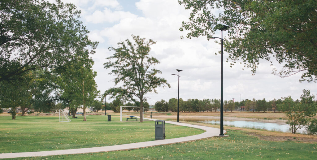 Curved walking path through a green park lined with Fonroche solar streetlights, with trees, a soccer field, and a small lake visible under a partly cloudy sky.
