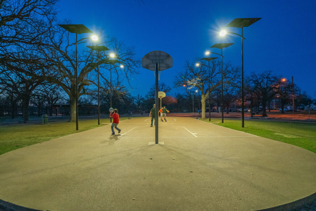 Children playing on a well-lit outdoor basketball court at dusk, illuminated by Fonroche solar-powered lighting systems in a tree-lined public park.