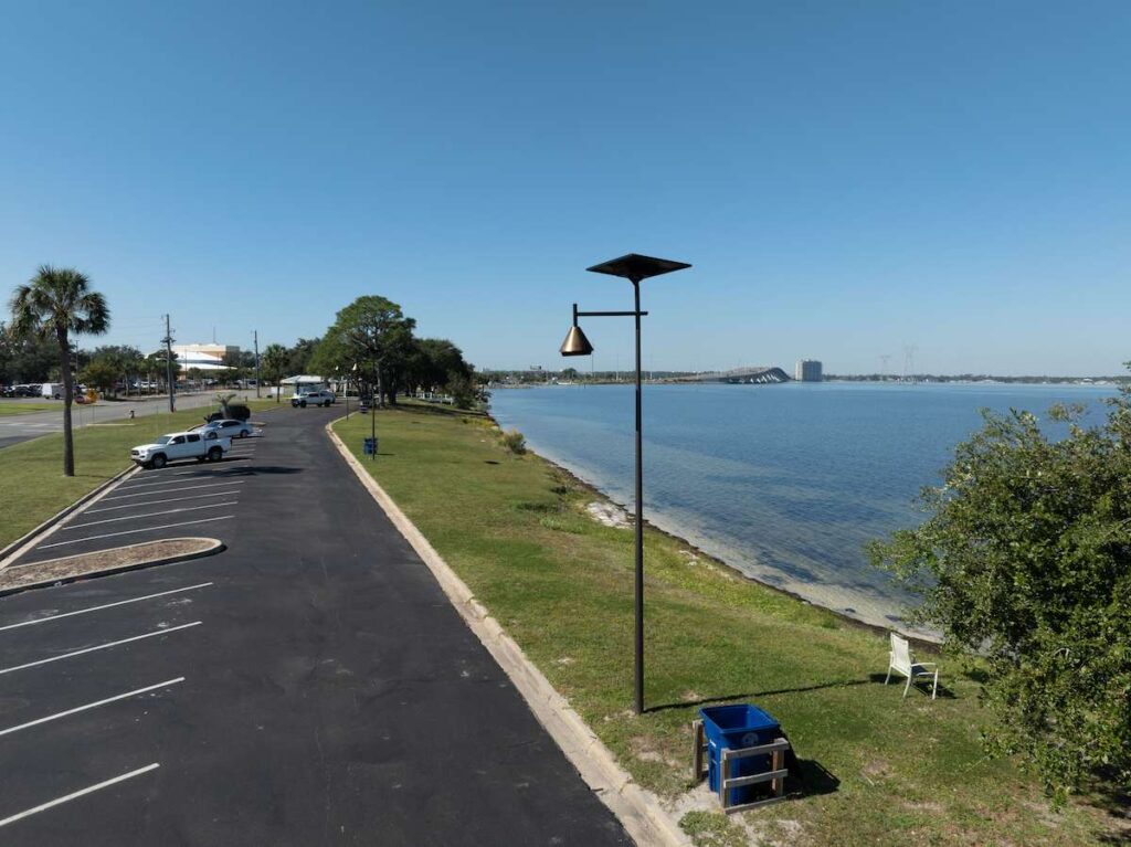 A solar-powered Fonroche streetlight illuminates a coastal waterfront parking area with palm trees and shoreline, ideal for hurricane-prone regions like coastal South Carolina.