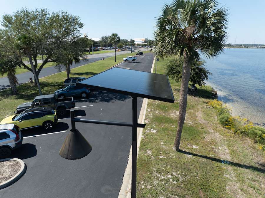 A close-up of a solar-powered streetlight in a coastal South Carolina-style waterfront parking area, with palm trees, blue sky, and calm shoreline.