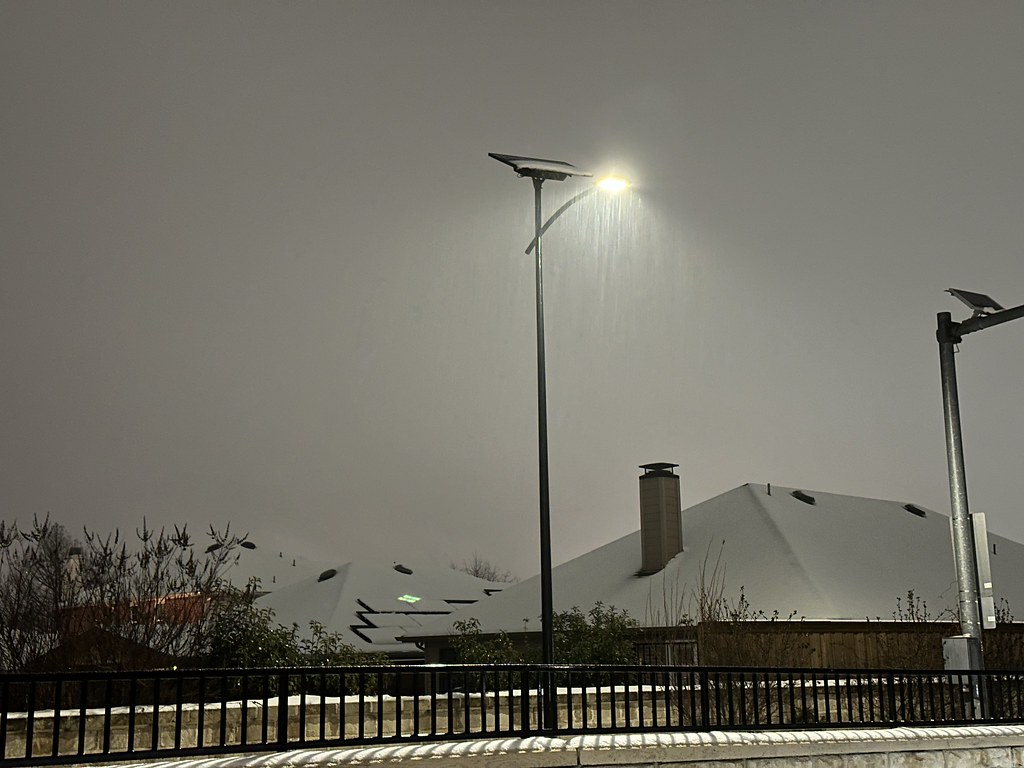 Fonroche solar street light shining through falling snow at night in a residential neighborhood, with rooftops and trees covered in a light layer of snow.