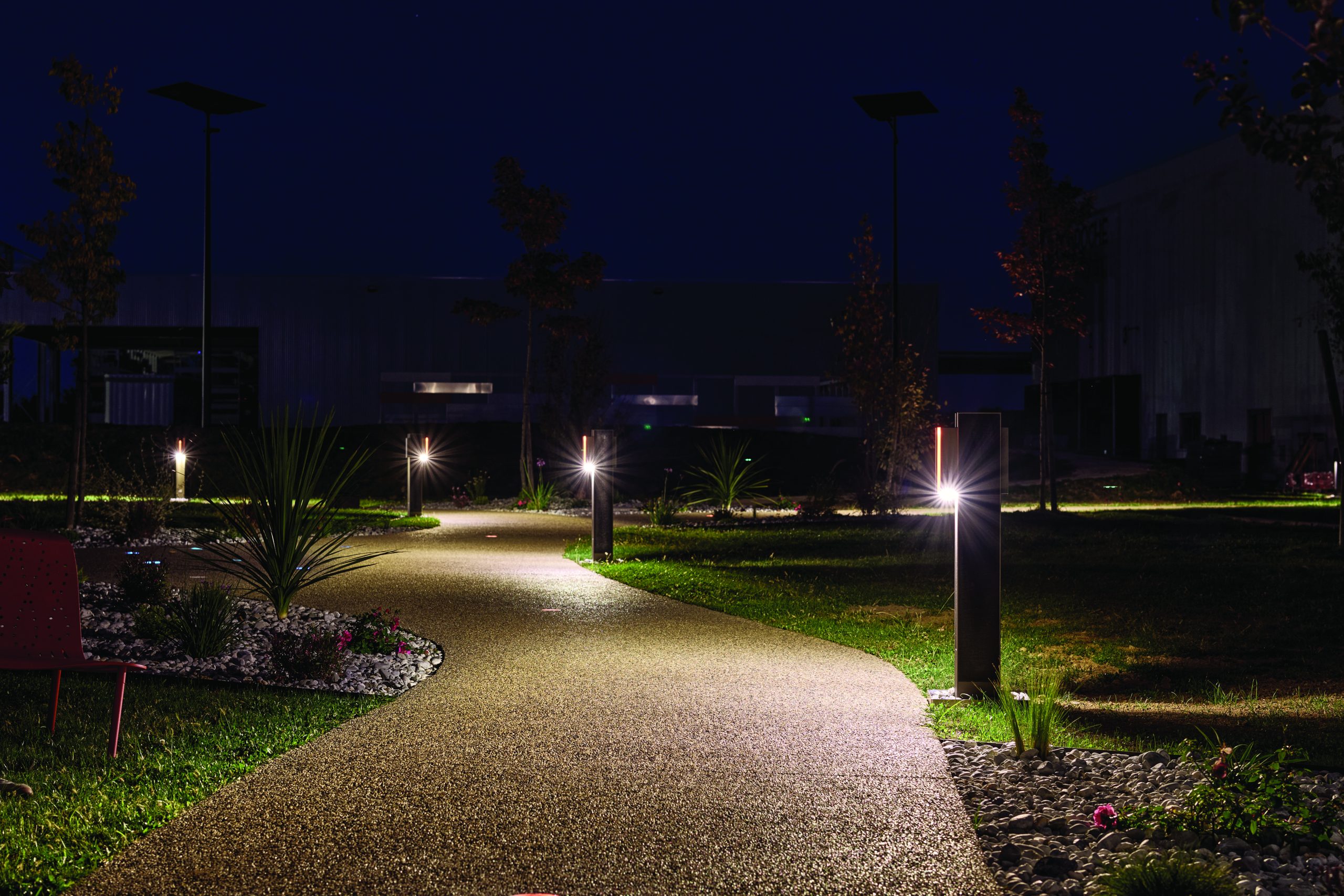 FOLLOW solar bollard pathway lights illuminating a landscaped pedestrian walkway at night in a modern park environment with soft, architectural lighting.
