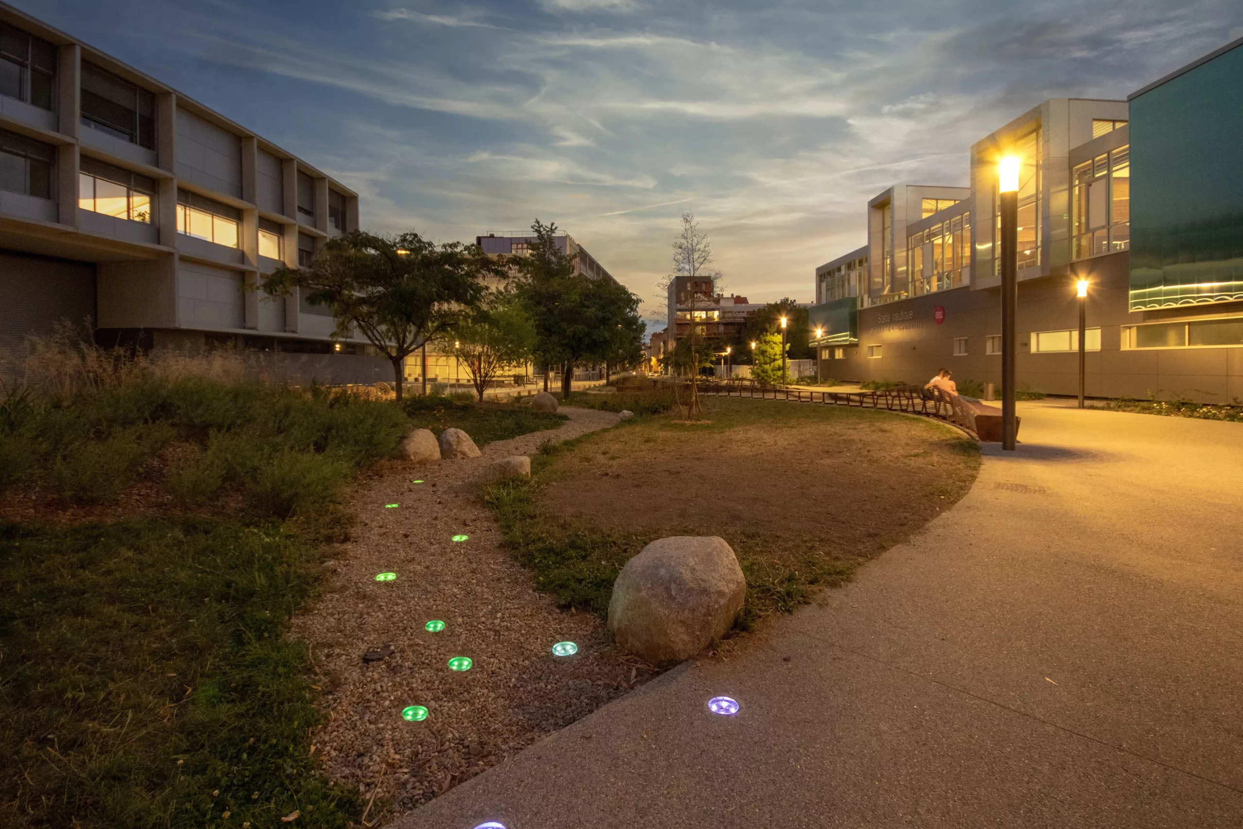 Colored illuminated pathway studs following a gravel garden path beside planting, rocks, and a paved courtyard at dusk.