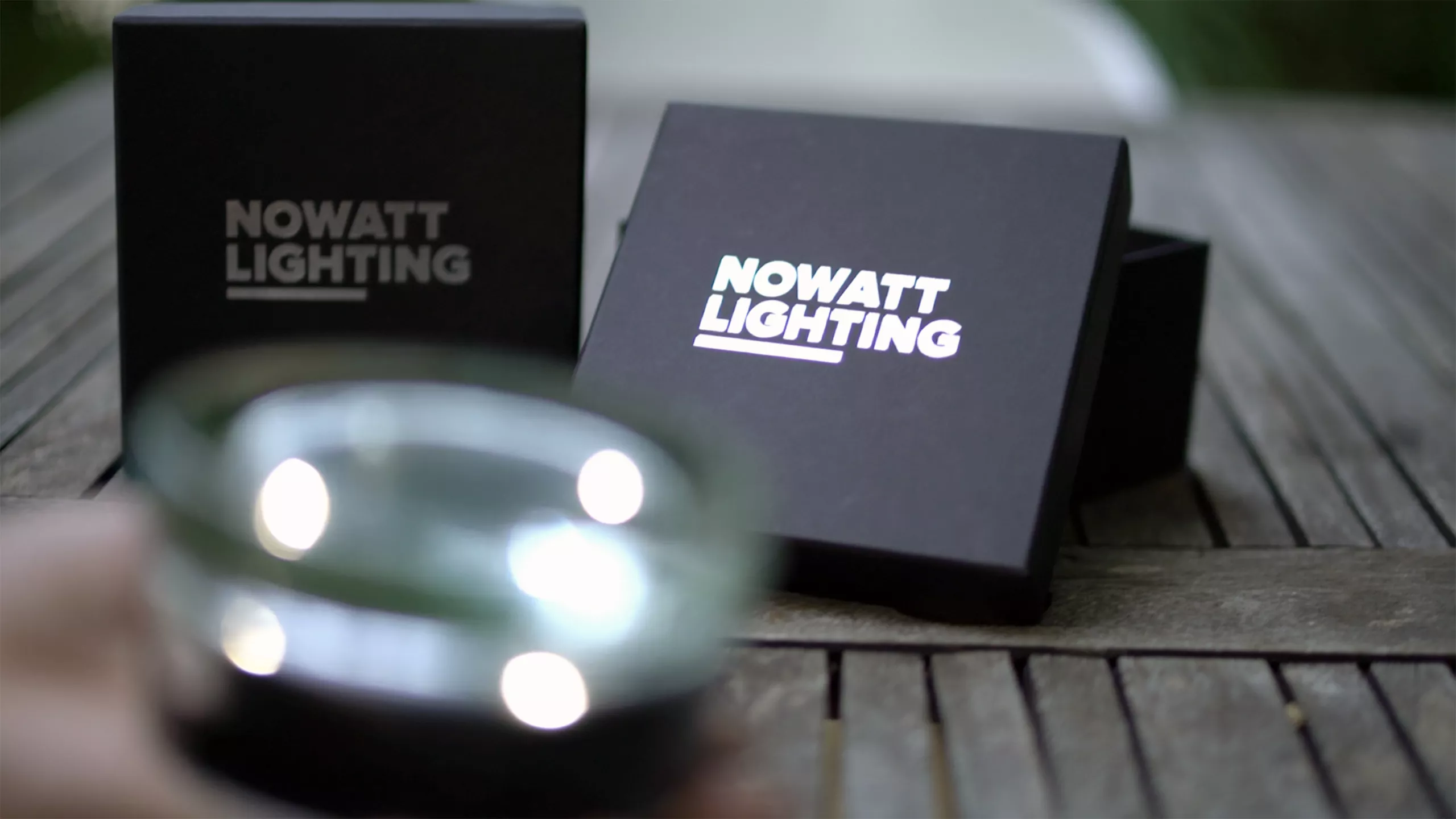 Close-up of an illuminated NOWATT Crystal Stud with branded presentation boxes on a wooden table.