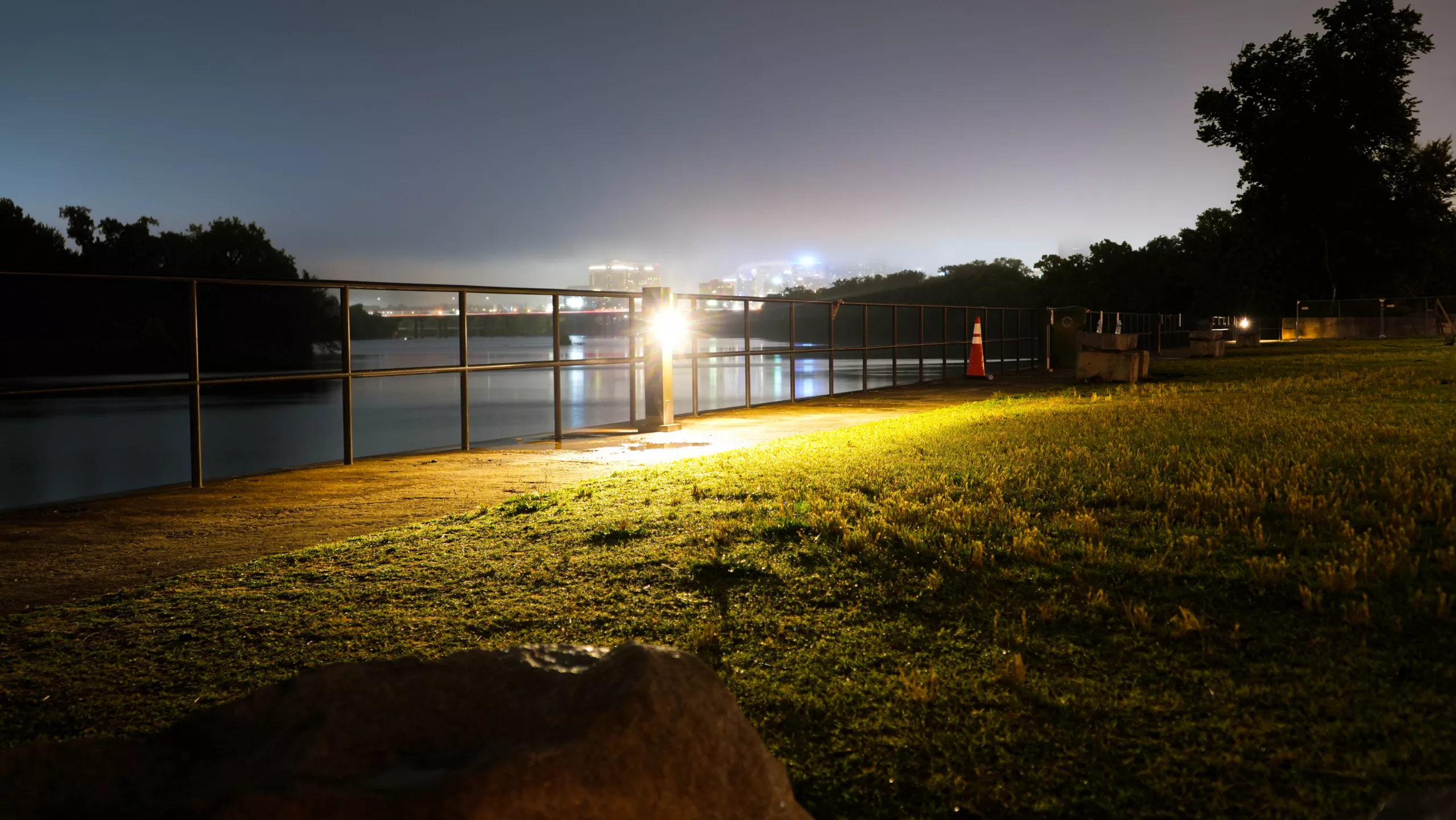 Bollard lights illuminating a waterfront walkway beside a metal railing and grass at night.