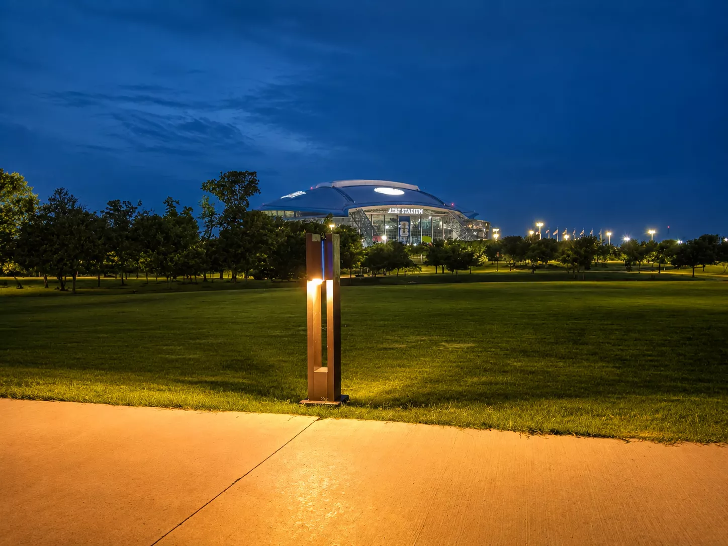 Illuminated bollard beside a concrete pedestrian path with AT&T Stadium in the background.