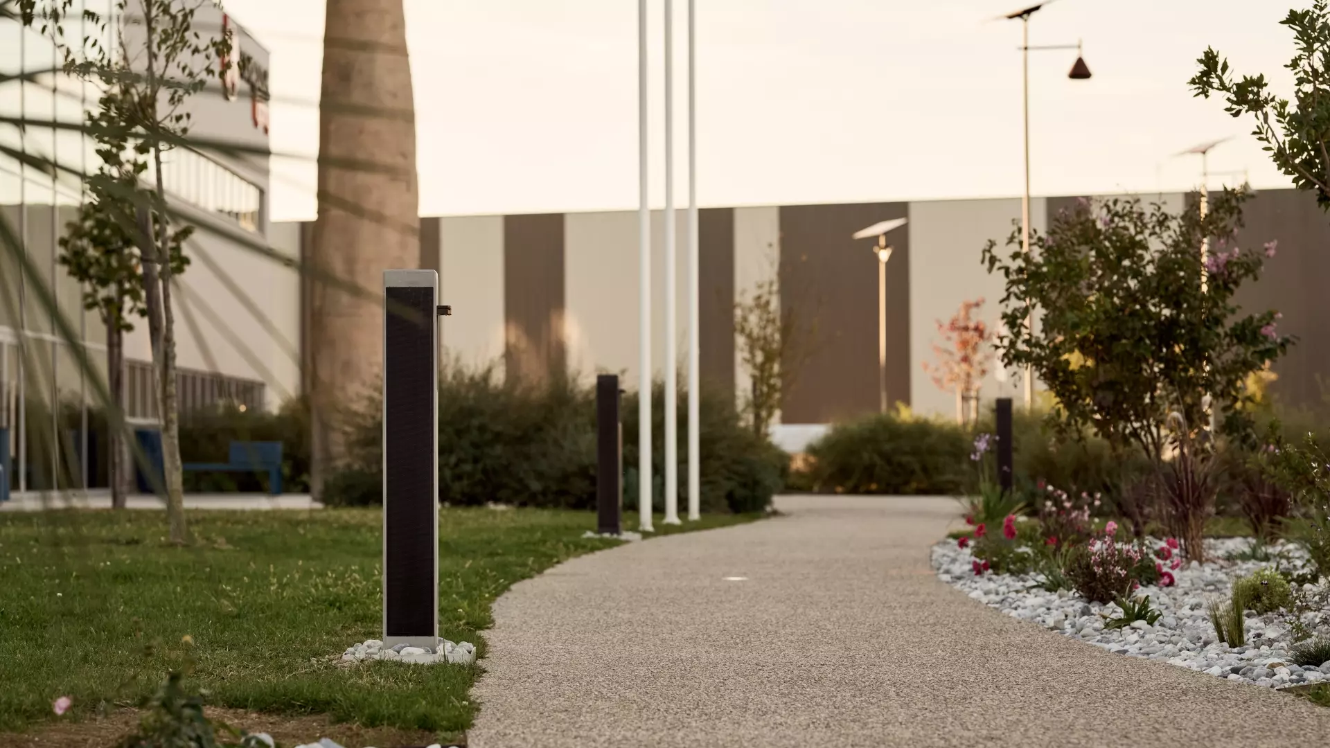 Solar bollards lining a curved courtyard walkway beside lawn, flowering plants, and buildings.