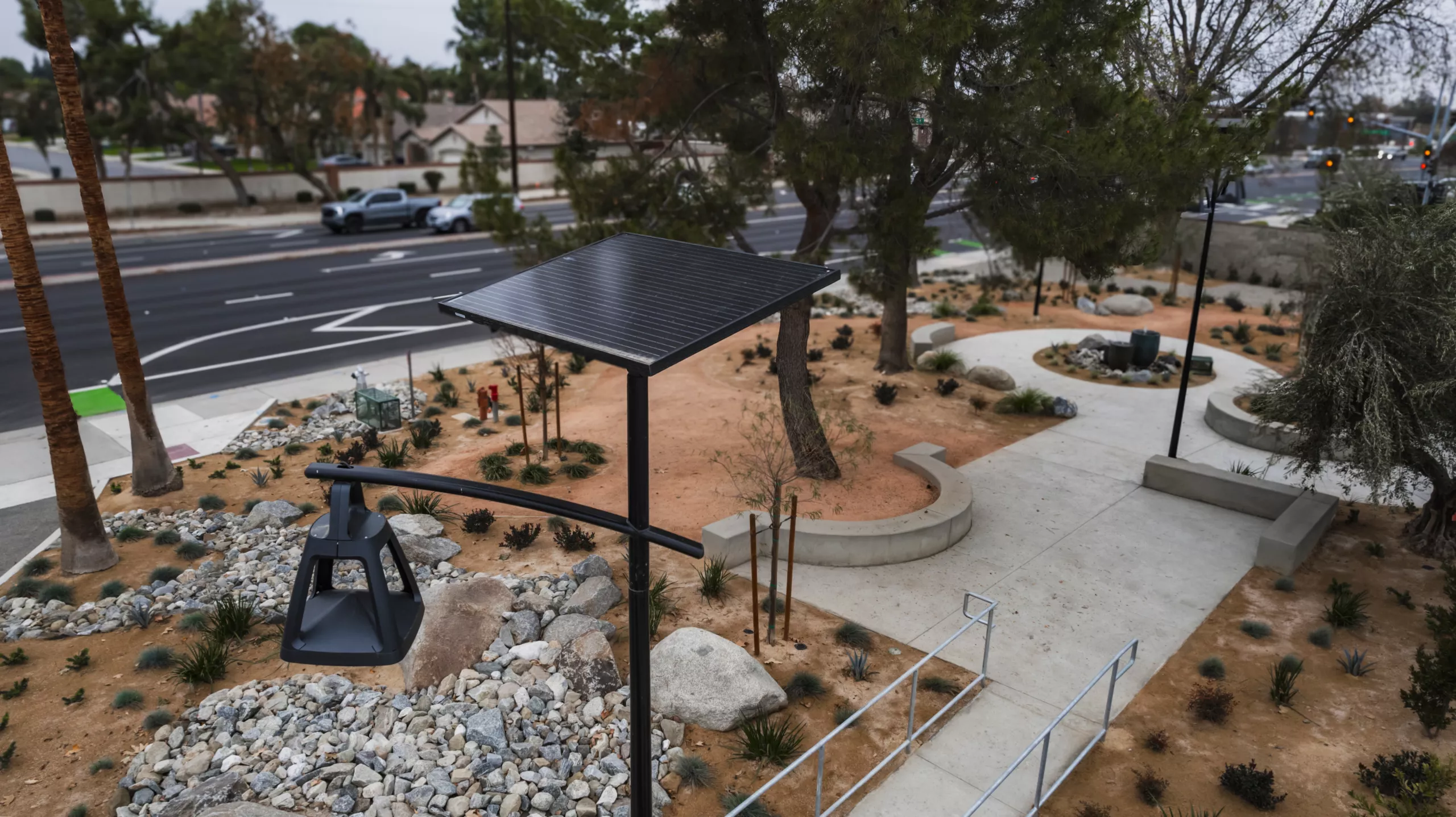 Close-up of a pole-mounted solar panel and light fixture beside a landscaped pathway and trees.